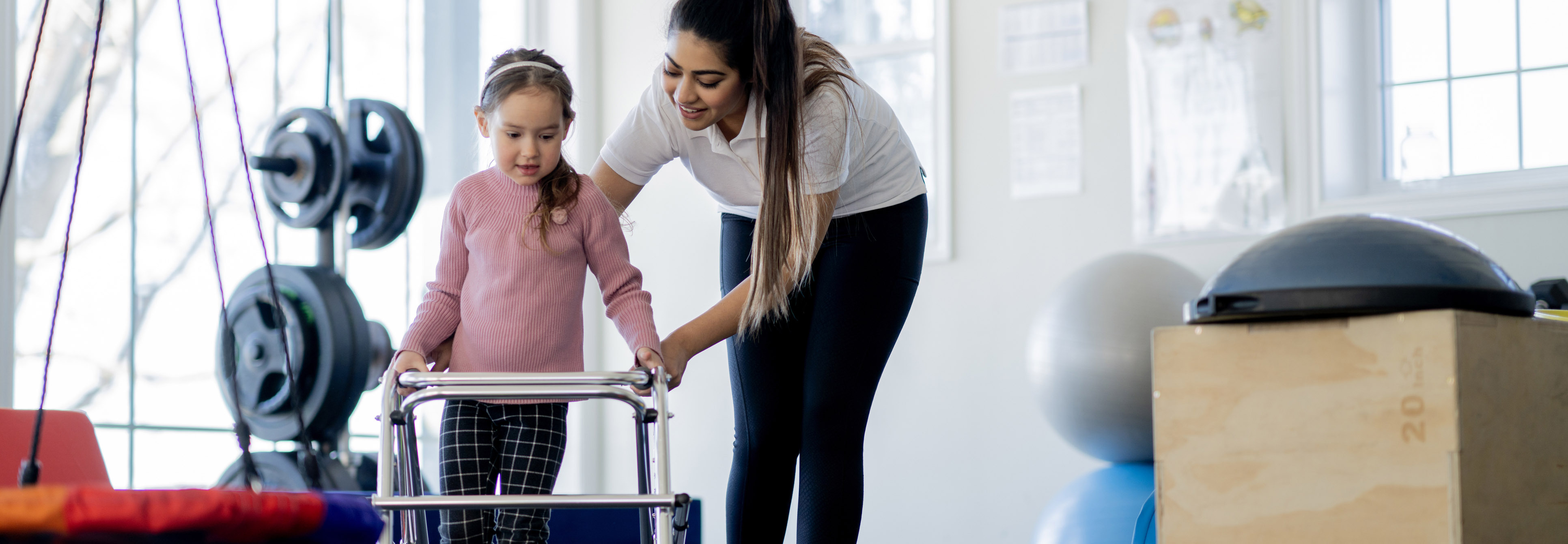 Young girl and adult in physical therapy walking with a walker.